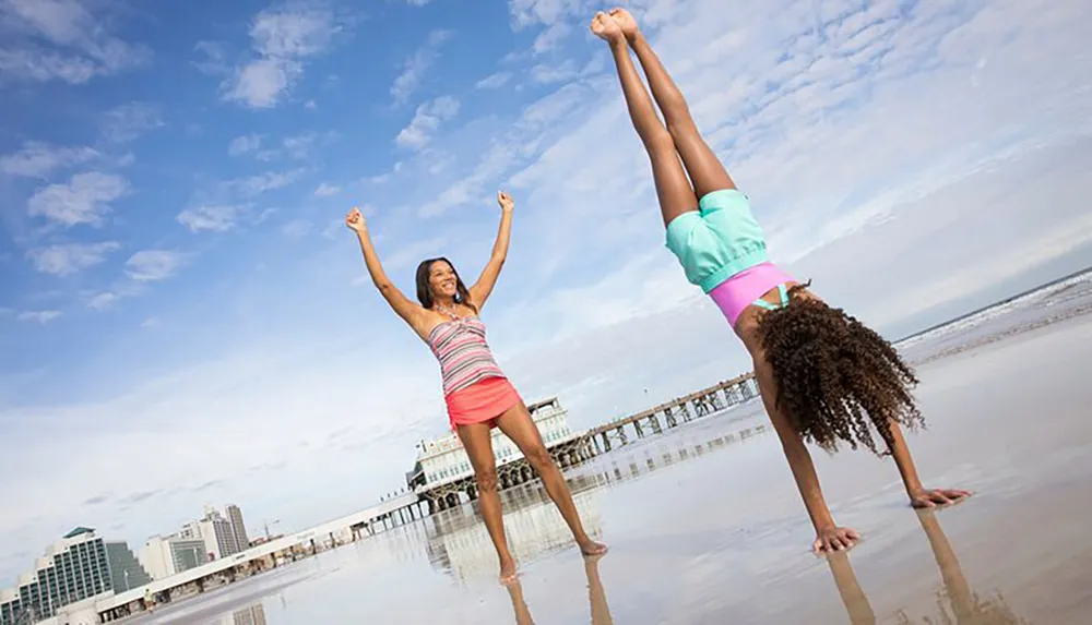 Two people enjoy a playful moment on a beach with one standing upright cheering and the other performing a handstand