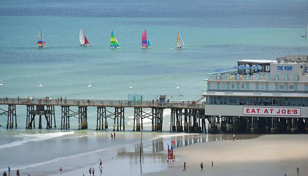 The image shows a bustling beach scene with a wooden pier featuring a restaurant and several colorful sailboats on the water in the background