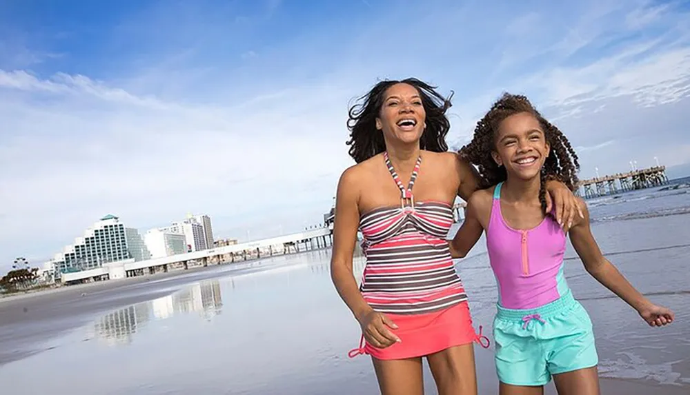 A joyful woman and a young girl are running on a sunny beach with a pier and buildings in the background both smiling and looking very happy