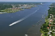 The image shows an aerial view of a wide river with a variety of boats and a cityscape in the background, creating a lively scene of waterway transportation and urban life.