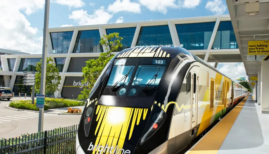 A colorful Brightline train is parked at a station on a sunny day with modern architecture in the background.
