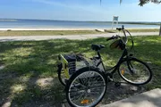 A three-wheeled bicycle with a basket and a hat is parked by a waterway on a sunny day.