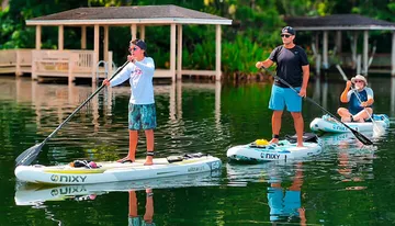 Three individuals are enjoying paddleboarding on a calm waterway, with lush greenery in the background and a dock to one side.