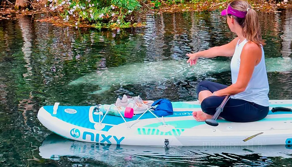 A person sits cross-legged on an inflatable paddleboard floating on a clear body of water holding a paddle and touching the water surface with their hand