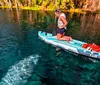 A person is standing on a paddleboard over clear water revealing underwater features with lush greenery in the background