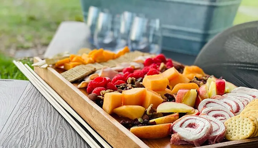 A charcuterie board filled with a variety of cheeses, fruits, nuts, and meats is presented on an outdoor table with wine glasses and a bucket in the background.