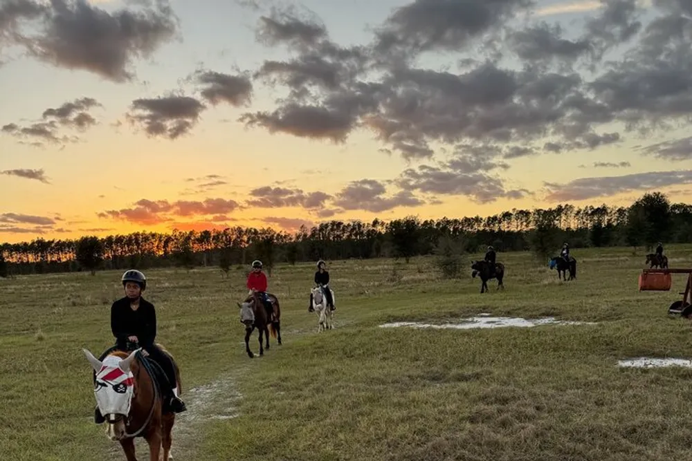 A group of riders on horseback enjoy a tranquil ride in an open field against the backdrop of a beautiful sunset