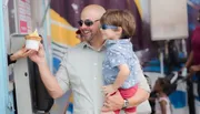 A smiling man carrying a young child is accepting an ice cream cone from a vendor at an outdoor event.