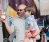 A smiling man wearing sunglasses is holding a child who is also wearing sunglasses as they seem to enjoy an outing together possibly getting some ice cream