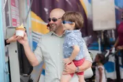 A smiling man wearing sunglasses is holding a child who is also wearing sunglasses, as they seem to enjoy an outing together, possibly getting some ice cream.