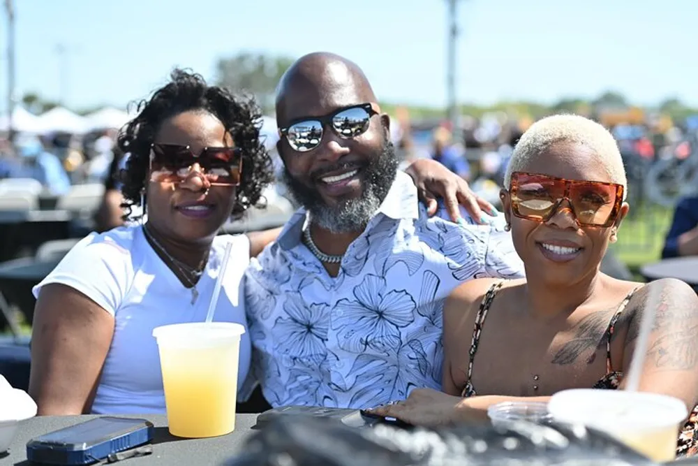 Three smiling adults are wearing sunglasses and enjoying an outdoor event together