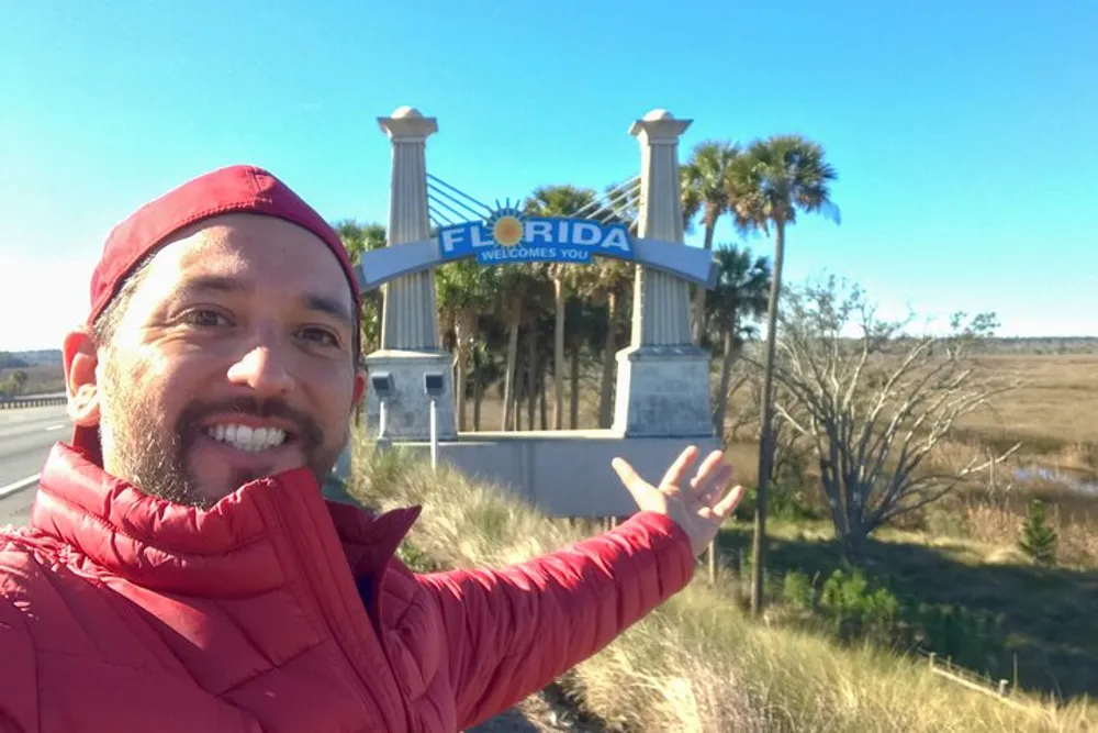 A person is taking a selfie in front of a Florida Welcomes You sign indicating they have just entered the state of Florida