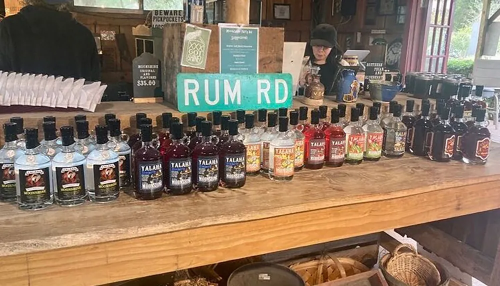 A variety of colorful bottles of spirits are on display at a rustic wooden counter under a sign reading RUM RD with a person working behind the counter