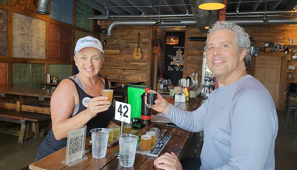 Two people are smiling and toasting with small glasses of beer at a rustic bar with number 42 on their table