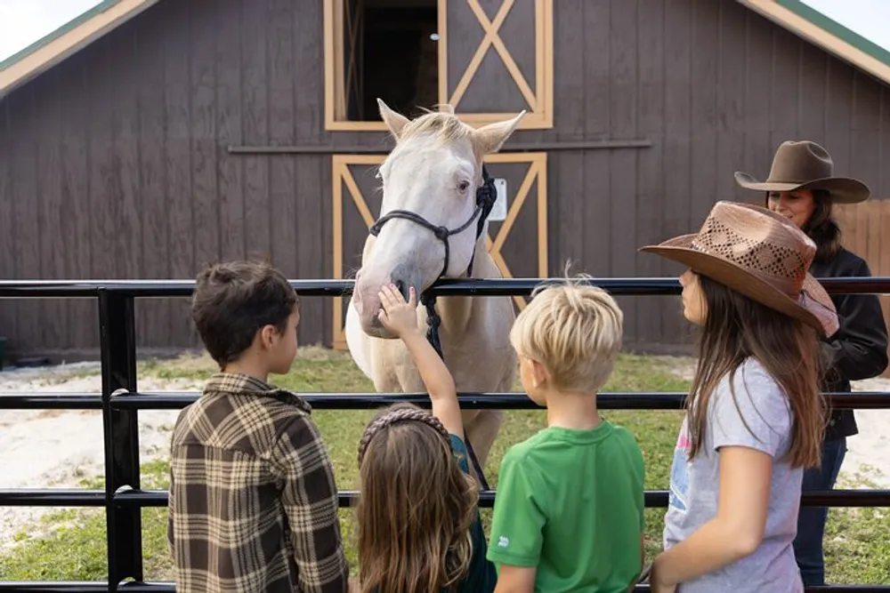 A group of children with one wearing a cowboy hat are petting and admiring a light-colored horse at a farm with an adult supervising