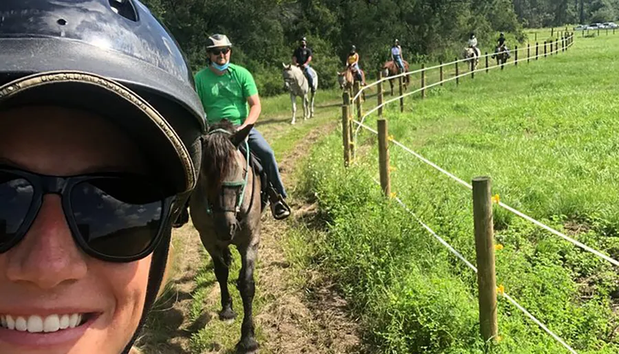 A group of people enjoying a horseback riding session on a sunny day, captured in a selfie by a rider at the front with her helmet visible.