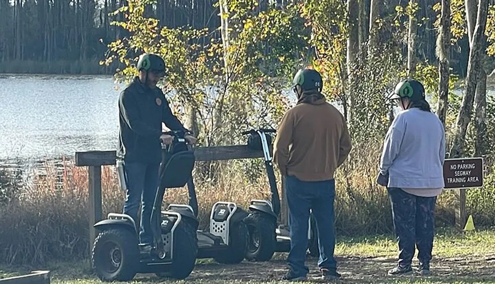 Three individuals wearing helmets are standing near a body of water with two Segway personal transporters by a sign indicating a No Parking Segway Training Area