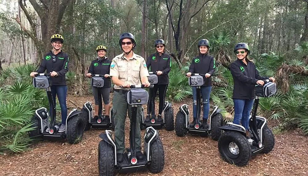 A group of six individuals wearing helmets and similar jackets is smiling for the camera while standing on Segway personal transporters in a forested area