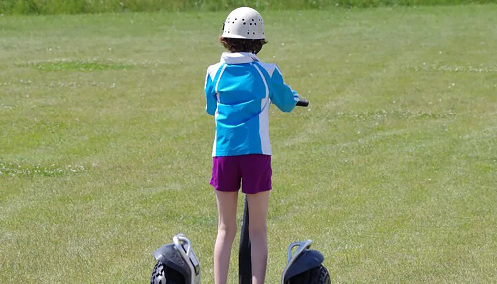 A person in a blue and white jacket and purple shorts is standing on a green grassy field wearing a helmet and preparing to ride a two-wheeled self-balancing personal transporter