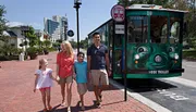 A family walks hand in hand on a sunny day next to a green trolley showing a sign that reads 