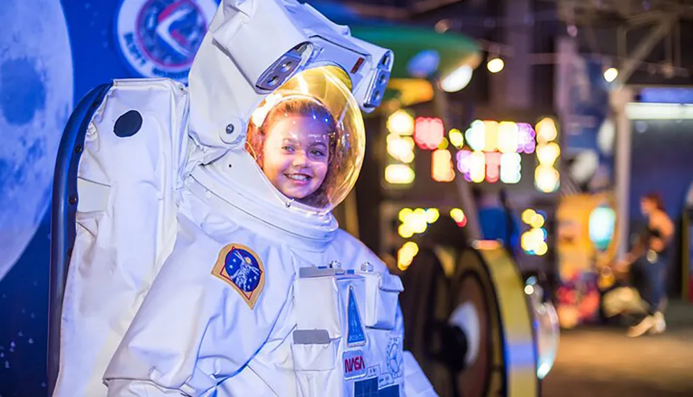 A person is seen smiling inside an astronaut suit at a location featuring space-themed dcor and colorful background lighting