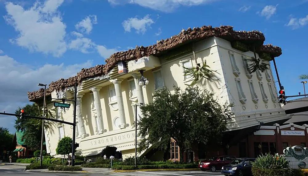 The image shows a building designed to look as though it is upside down with its roof on the ground and the base in the air under a blue sky with scattered clouds