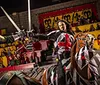 The image depicts a staged medieval jousting scene between two armored individuals on horseback in front of an audience alongside an inset photo of roasted poultry with accompaniments representing a themed dining experience