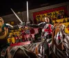 Two knights on horseback are engaged in a staged sword fight at a medieval-themed tournament as spectators watch from the stands