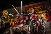 Two knights on horseback are engaged in a staged sword fight at a medieval-themed tournament as spectators watch from the stands.