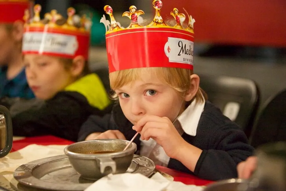 A child wearing a paper crown with a medieval theme is drinking soup through a straw at a festive event