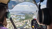 Two people are piloting a helicopter over a scenic coastal area with a clear view of the landscape and waterways below.