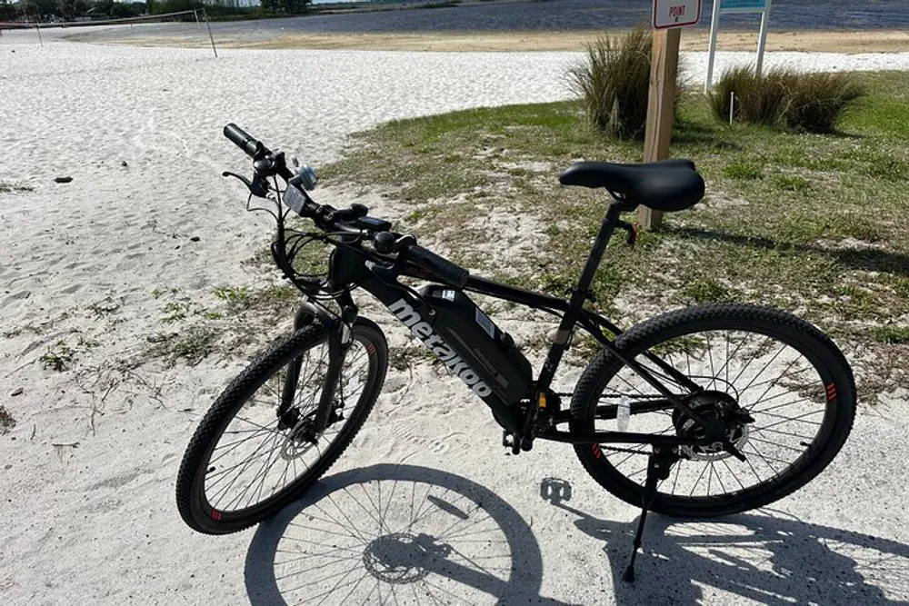 A black electric bicycle is parked on a sandy area near a beach under sunny conditions