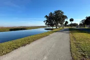 A serene pathway runs alongside a waterway, flanked by green grass and trees under a clear blue sky.