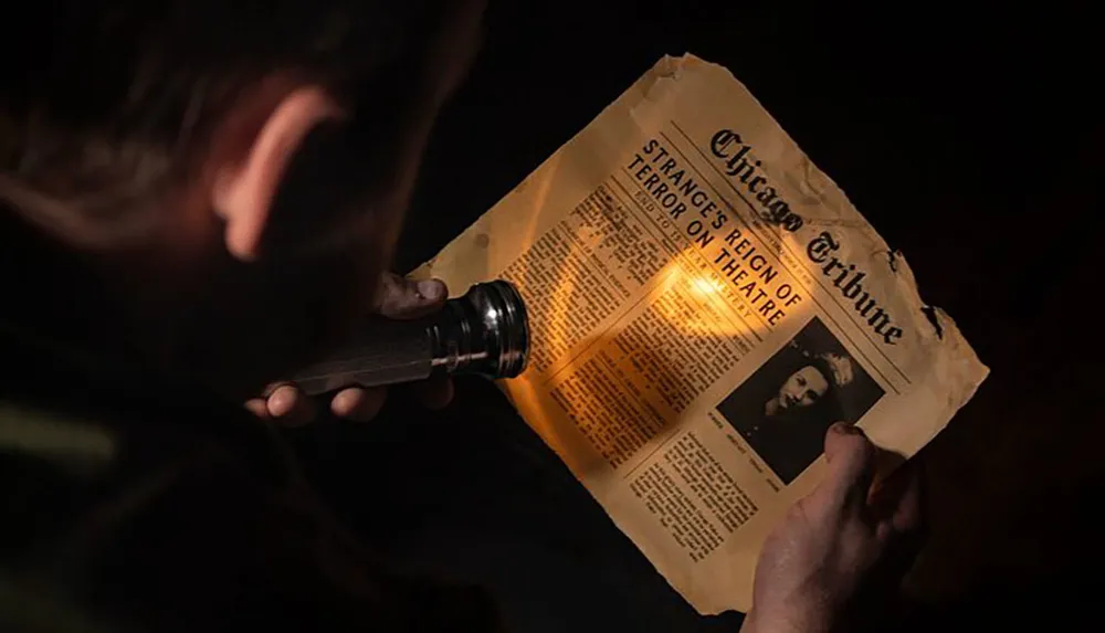 A person is holding an old torn newspaper article from the Chicago Tribune with a headline about terror at a theatre illuminated by a flashlight in a dimly lit setting