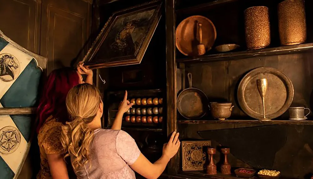 Two individuals are examining or searching for something in a rustic wooden cabinet filled with assorted antique kitchenware and decorations