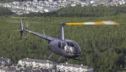 A helicopter flies over a forested area near a housing development, its rotor blades highlighted by the sun to create an illusion of a discontinuous appearance.