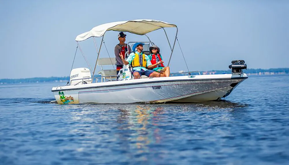 Three people wearing life jackets are enjoying a sunny day on a small motorboat with a Bimini top on a calm water body