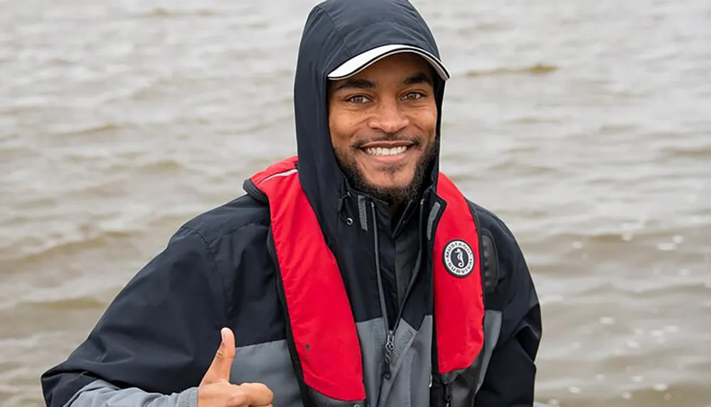 The image features a smiling individual giving a thumbs-up while wearing a hooded jacket and life vest with a water body in the background