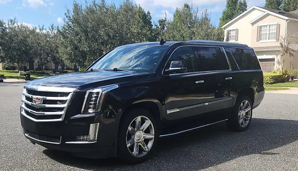 A black Cadillac Escalade SUV is parked on a suburban street in front of a house under a clear sky