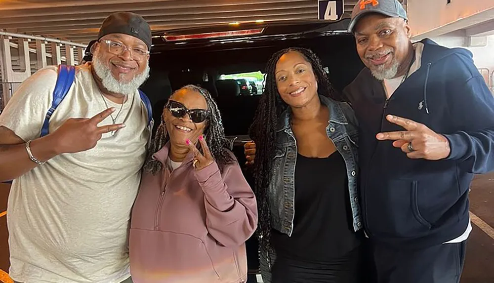 Four adults are smiling and posing for a photo with two of them making peace signs with their hands