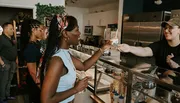 A smiling customer is receiving a cup of ice cream from a friendly server at an ice cream shop, with other customers waiting in line behind her.