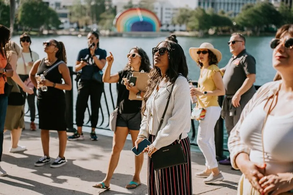 A group of people are enjoying a sunny day near a body of water with a colorful rainbow structure in the background