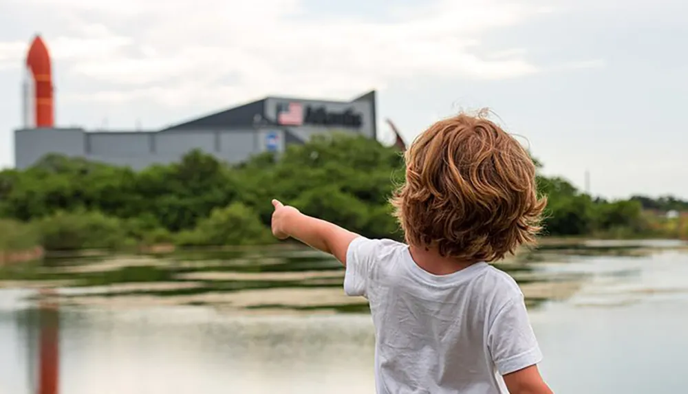 A child with curly hair is pointing towards the Space Shuttle exhibit at the Kennedy Space Center as seen across a body of water