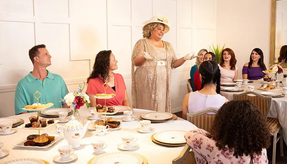 A group of people is enjoying a festive tea party with a standing woman in a vintage dress and hat likely entertaining or hosting the event