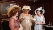 Three women dressed in vintage clothing with elaborate hats are having a cheerful conversation in an elegantly furnished room.