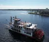 This image features a large red and white paddlewheel riverboat docked beside a calm body of water under a clear blue sky