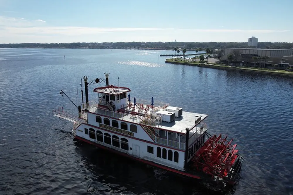 A paddlewheel boat is cruising on a calm river with a clear sky overhead and a scenic shoreline in the background