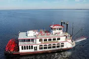 This image features a large red and white paddlewheel riverboat docked beside a calm body of water under a clear blue sky.