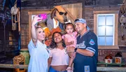 A family is taking a selfie with a person in an oversized dog mascot costume wearing sunglasses in a rustic, wood-paneled room.