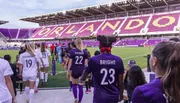 Players are walking onto a soccer pitch in a stadium with 'ORLANDO' displayed across the seating.